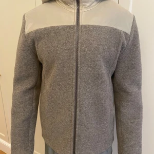 Grå hybridjacka från Colmar - Snygg grå jacka från Colmar med ljusgrå detaljer på axlar, huva och armbågar. Jackan har hel dragkedja framtill, huva och raka linjer. Materialmix med ull och syntet ger en modern look. Perfekt för dig som gillar stilren design och vill ha något unikt.