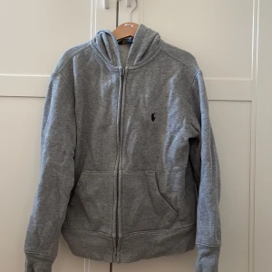 Grå hoodie från Polo Ralph Lauren - Säljer en grå hoodie från Polo Ralph Lauren i storlek 122/128 med klassisk logga broderad på bröstet. Tröjan har huva, dragkedja framtill och två stora fickor. Perfekt för hösten! 