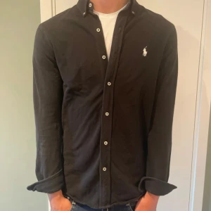 Svart skjorta från Ralph Lauren - Svart långärmad skjorta från Ralph Lauren med klassisk krage och vita knappar. Diskret broderad logga på bröstet. Tillverkad i lätt bomullsmaterial (featherweight mesh) som känns skön mot huden. Perfekt för dig som gillar stilrena och tidlösa plagg.