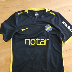 AIK svart fotbollströja Nike #18 Medium (small) - Står medium men passar small mer.                      AIK fotbollströja från Nike med korta ärmar, svart bas och gula detaljer. Tryck med sponsorer, klubbmärke på bröstet och nummer 18 på ryggen. Materialet är lätt och andas, perfekt för match eller träning. Snygg gul Nike-logga och gula inslag vid krage och ärmslut.