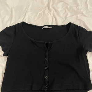 Svart croppad topp från 157 - Svart croppad topp från 157 med korta ärmar och ribbad struktur. Toppen har rundad halsringning och knappar framtill som ger en chill vibe. Perfekt för att styla med höga jeans eller kjol. Materialet är mjukt och stretchigt, vilket gör den bekväm att bära.