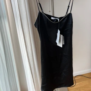 Svart kort klänning med strass från NA-KD - Supersnygg svart kort klänning från NA-KD med tunna axelband och strassdetaljer längs halsringningen. Klänningen har en slät satinliknande yta som ger en lyxig känsla och är perfekt för dig som vill sticka ut. Stilren och trendig design.