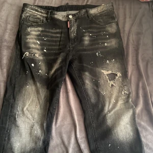 Svarta jeans från Dsquared2 med slitningar - Snygga svarta jeans från Dsquared2 med coola slitningar och färgstänk för en edgy vibe. Jeansen har klassisk femficksdesign, läderpatch bak och en tvättad look med ljusare partier. Perfekta för dig som gillar streetstyle och vill sticka ut. Storlek 38!