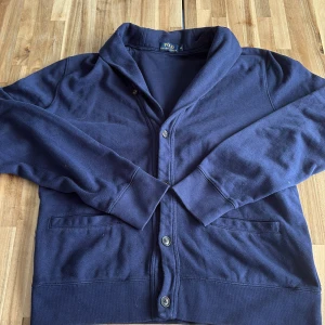 Mörkblå kofta från Polo Ralph Lauren - Snygg mörkblå kofta från Polo Ralph Lauren med klassisk krage och knappar framtill. Tröjan har två fickor och är långärmad, perfekt för att ge en chill och stilren vibe. Materialet är mjuk bomull som känns skönt mot huden. Den är i jätte fint skick och passar dej som har M/S. Ny pris 2200 mitt pris 450