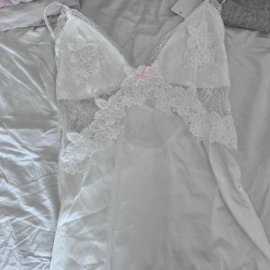 Vit spetsklänning / nattlinne lingerie med rosett - Supersöt vit kortklänning/nattlinne, lingerie med tunna axelband och transparent spets upptill. Blommiga spetsdetaljer och en liten ljusrosa rosett framtill ger en romantisk vibe. Klänningen har omlottkänsla vid bysten och är tillverkad i mjukt, lätt material som känns lyxigt mot huden NY MED PRISLAPP. Köptes för 400kr
