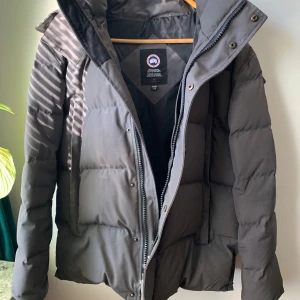 Grå dunjacka från Canada Goose - Grå canada goose wyndham storlek large, fint skick. Finns en defekt inne i ena fickan, ett hål men annars felfri . Pris 3500kr