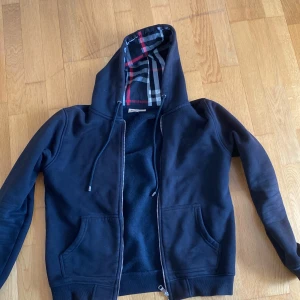 Burberry kofta - Svart hoodie från Burberry med klassiskt rutigt foder i huvan. Tröjan har dragkedja framtill, snörning vid huvan och två fickor. Materialet är mjuk bomull och passformen är normal. Perfekt för dig som gillar stilrena detaljer och ikoniskt mönster.