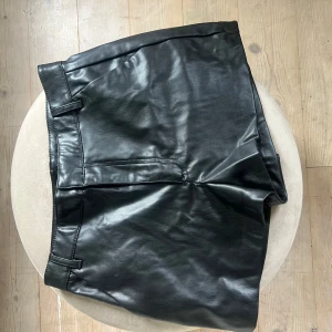 Svarta skinnshorts från Zara - Snygga svarta shorts från Zara i skinnimitation med klassisk rak passform och midwaist. De har bälteshällor och diskreta fickor fram, vilket ger en clean och stilren look. 