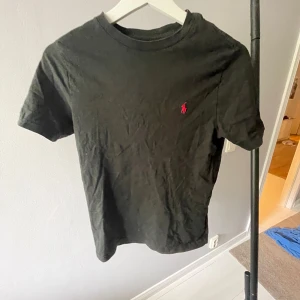 Svart t-shirt från Polo Ralph Lauren - Svart t-shirt från Polo Ralph Lauren med klassisk röd logobrodyr på bröstet. Modellen är kortärmad och har en rund halsringning. Tillverkad i mjuk bomull som är skön mot huden. Perfekt för en clean och enkel stil.140/152