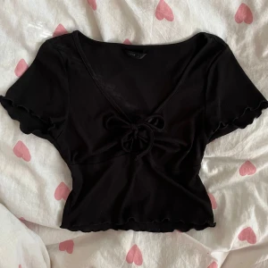 Svart croppad topp med knytning - Svart croppad topp från SHEIN i strl XS med ribbad struktur och v-ringning. Toppen har korta ärmar med vågig kant och en söt knytning framtill!💞                                                                OBS! Säljer en i ljusgrön också