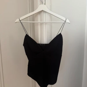 Svart body med smala axelband - Svart linnebody från Nelly med smala axelband och v-ringad front. Toppen har en stilren och enkel design som är lätt att matcha med olika outfits. Materialet känns mjukt och lätt mot huden, perfekt för varma dagar eller när du vill ha en clean look.