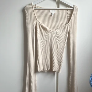 Ribbad beige långärmad topp H&M - Säljer en beige ribbad topp från H&M med lång ärm och snygg v-ringad hals. Toppen har en figurnära passform och coola sömdetaljer framtill som ger en modern vibe. Perfekt att styla med jeans eller kjol för en trendig look.