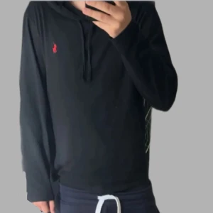 Svart hoodie från Polo Ralph Lauren - Svart hoodie från Polo Ralph Lauren med klassisk röd broderad logga på bröstet. Tröjan har huva med snörning och är tillverkad i mjuk bomull. Långärmad och perfekt för en chill och stilren look.