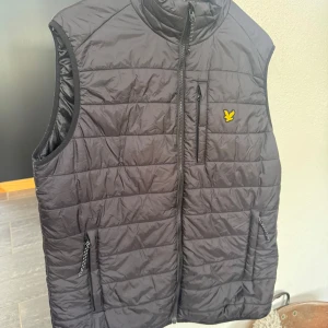 Svart dunväst från Lyle & Scott - Riktigy snygg svart dunväst från Lyle & Scott med loggan på bröstet. Västen har quiltad design, krage och dragkedja framtill. Två sidofickor med dragkedja och en bröstficka. Mycket trendig väst perfekt till lager på lager och uppklädda tillfällen eller att slänga över en hoodie för extra värme och en casual look. Västen är bara använd 1 gång tidigare och nästintill helt i nyskick förutom att lappen inte är kvar. Ordinarie pris låg på 1000, pris kan diskuteras