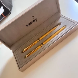 Pennset - Pris kan diskuteras,Bilderna visar ett exklusivt set med två guldfärgade pennor i en elegant ask med texten 'Melody'. Detta är inte en bok, utan ett pennset som passar perfekt för dig som vill skriva med stil, anteckna eller ge bort som present.