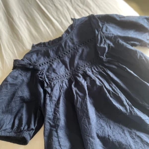 Marinblå top från Gina Tricot - Supersöt marinblå topp från Gina Tricot med korta puffärmar och volangdetaljer vid axlarna. Smockad överdel med snygga broderade detaljer och luftig passform. Perfekt för dig som gillar en chill och trendig look.