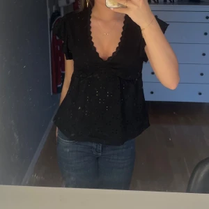 Svart blus med v-ringning och spetsdetaljer - Säljer en svart blus med djup v-ringning och fina spetsdetaljer längs urringningen. Blusen har korta ärmar och lätt volang vid axlarna. Perfekt att styla med jeans eller kjol.