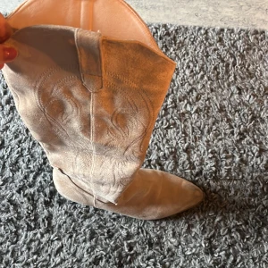 Beiga cowboy boots i mocka - Snygga beiga cowboy boots i mocka med klassiska broderade detaljer och spetsig tå. Skorna har en låg klack och högt skaft, perfekta för att lyfta din outfit med western-vibe. Står stl 41 men jag har själv 38,5 ..