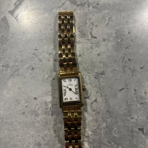 Guldfärgad klocka från Mockberg - Säljer en elegant guldfärgad klocka från Mockberg med rektangulär vit urtavla och romerska siffror. Klockan har ett klassiskt länkarmband i metall och en stilren design som passar till många tillfällen. Skriv för fler bilder eller liknande