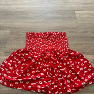 Röd blommig kjol med smock - Supersöt röd kjol med vitt blommönster och bred smock i midjan. Kjolen har en luftig och svängande passform, perfekt för varma dagar. Den är kort och har en härlig sommarkänsla.