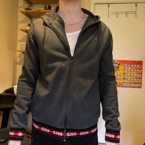 Mörkgrå hoodie med dragkedja från Hugo boss junior märke Hugo  - Säljer en mörkgrå hoodie med dragkedja från Hugo. Jackan har huva med snörning och ribbade muddar. Detaljerad med röd och vit logotyp längs nederkant och ärmslut. Perfekt för en avslappnad och stilren look.