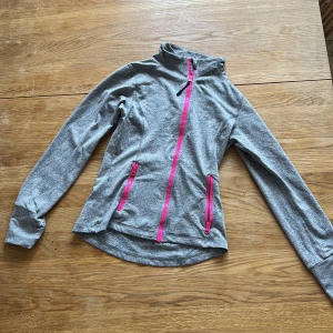 Grå träningsjacka med rosa dragkedjor - Säljer en grå träningsjacka med snygga rosa dragkedjor och hög krage. Jackan har två fickor framtill och är tillverkad i ett stretchigt syntetmaterial. Perfekt för utomhusaktiviteter eller gymmet. Den är tight din mage. Väldigt fin men har en ny💕💕