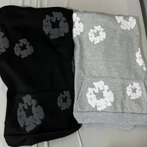 Svart och grå hoodie med blommigt mönster - Två hoodies i svart och grått med stora blommiga tryck i vitt och grått. Båda har huva och magficka, samt ribbade muddar vid ärmslut och nederkant. Perfekt för en avslappnad stil.