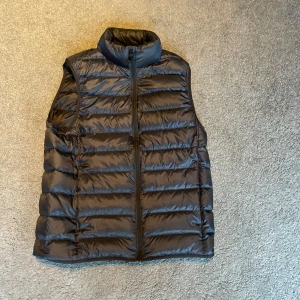 Svart dunväst från Uniqlo - Säljer en svart dunväst från Uniqlo i storlek XS. Västen har quiltad design, hög krage och dragkedja framtill. Perfekt för lager-på-lager och lätt att bära tack vare sitt tunna och smidiga material.