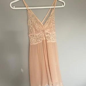 Ljusrosa nattlinne från Hunkemöller - Säljer en ljusrosa nattlinne från Hunkemöller med spetsdetaljer och smala axelband. Nattlinnet har v-ringning, transparent mesh med små prickar och korsade band i ryggen. Perfekt för dig som gillar romantisk och feminin stil.