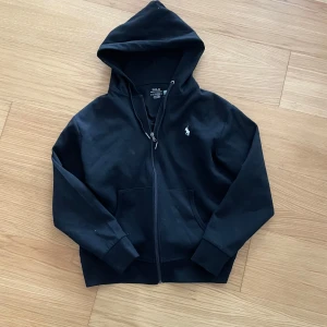 Zip hoodie från Ralph Lauren - Säljer en svart hoodie från Polo Ralph Lauren med dragkedja och huva. Tröjan har två fickor framtill och en liten broderad logga på bröstet. Perfekt för en avslappnad och stilren look.