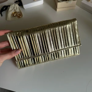 Guldig kuvertväska med kedja - Säljer en glansig guldig kuvertväska med räfflad yta och kedja som axelrem. Perfekt för att lyfta din outfit med lite extra bling. Väskan har ett stilrent och modernt uttryck. 💕
