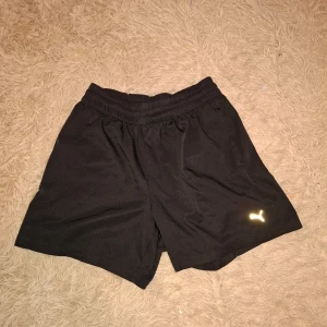Svarta träningsshorts från Puma - Svarta shorts från Puma med elastisk midja och klassisk logga i vitt på ena benet. Perfekta för träning eller sport. Lätta och bekväma med normal passform.