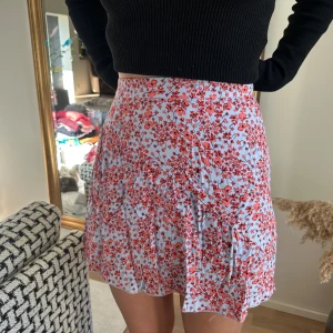 Vit blommig kortkjol med volang - Supersöt vit kjol med röda blommor och volangkant. Kjolen har en lätt utsvängd passform 