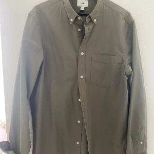 Olivgrön skjorta från H&M - Säljer en olivgrön skjorta från H&M i regular fit. Skjortan har långa ärmar, klassisk krage med knappar och en bröstficka. Perfekt för dig som gillar enkel och stilren design.