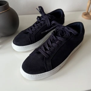 Mairinblåa sneakers från Les Deux i mocka - Snygga marinblåa sneakers från Les Deux i mocka med vita sulor. Skorna har snörning och en diskret detalj med röd och vit rand på hälen. Perfekta för en stilren look.