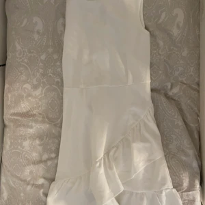 Vit ärmlös klänning med volang - Säljer en stilren vit klänning med ärmlös design och volangdetalj nedtill. Klänningen har en rak passform och är perfekt för sommarens alla tillfällen. Enkel och fräsch look som passar till många olika stilar.