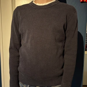 Mörkblå stickad tröja med knyt i sidan - Säljer en mörkblå stickad tröja med rund halsringning och långa ärmar. Tröjan har en ribbad struktur och en snygg knytdetalj i sidan vid midjan. Perfekt för dig som gillar enkel men stilren design.