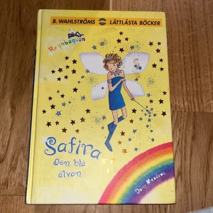 Safira den blå älvan - En barnbok från serien Regnbågsön, skriven av Daisy Meadows. Omslaget är gult med en tecknad älva i blå klänning, vingar och stjärnor runtom. Boken har hårda pärmar och färgglada detaljer, inklusive en regnbåge längst ner.