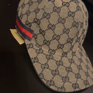 keps med monogram och röd-blå rand - Snygg keps med klassiskt GG-monogram i grått och beige. Kepsen har en röd och blå rand på sidan och är tillverkad i bomullsmaterial. Perfekt accessoar för en trendig look.