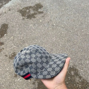 Gucci keps med monogram och röd-blå rand - Säljer en klassisk Gucci keps i grått med GG-monogram över hela kepsen och en röd-blå rand på sidan. Justerbar rem baktill för perfekt passform. Perfekt accessoar för dig som gillar exklusiva märken.