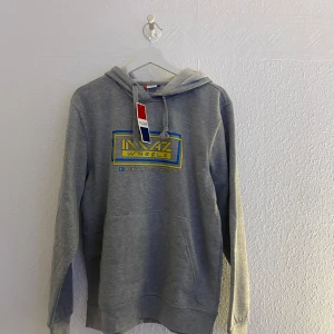 Grå hoodie från Clique med tryck - Säljer en grå hoodie från Clique med MAZ Wheels-tryck i gult och blått på bröstet och ärmen. Tröjan har huva med dragsko och en stor magficka. Perfekt för dig som gillar streetwear och snygga detaljer.