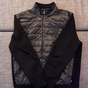 Svart hybridjacka från BOSS - Snygg svart hybridjacka från BOSS med quiltad framsida i glansig polyester och släta ärmar. Jackan har dragkedja framtill och diskret BOSS-logga på bröstet. Perfekt för dig som vill ha en stilren och modern look. Jag kommer från Tyskland och skickar även jackan från Tyskland. Du skulle behöva betala frakten själv och jag täcker inte det. Om du är intresserad, vänligen meddela mig.