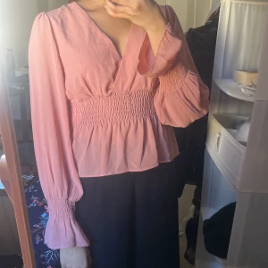 Rosa blus med smock från Amisu - Supersöt rosa blus från Amisu med v-ringning och smockad midja. Blusen har långa puffärmar med smock vid ärmslut och knappar framtill. Perfekt för dig som gillar romantiska detaljer och en feminin stil. Priset kan diskuteras :) 