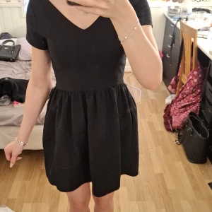 Svart kortklänning med v-ringning från Zara Trafaluc - Säljer en svart kortklänning från Zara Trafaluc med v-ringad hals och korta ärmar. Klänningen har en markerad midja och utställd kjol, vilket ger en klassisk och stilren look.