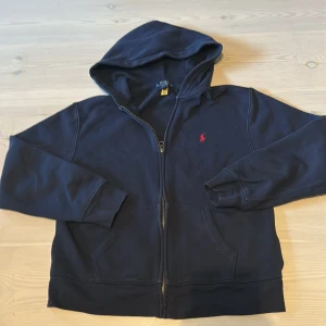 Mörkblå hoodie från Polo Ralph Lauren - Säljer en mörkblå ziphoodie från Polo Ralph Lauren med dragkedja och huva. Klassisk design med röd broderad logga på bröstet och två fickor framtill. Perfekt för en avslappnad stil. Nypris 1100kr