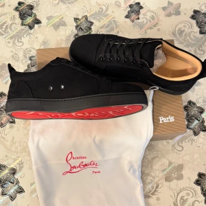 Svarta sneakers från Christian Louboutin med röd sula - Säljer ett par svarta sneakers från Christian Louboutin med ikonisk röd sula och diskret logga på sidan. Skorna har snörning, rund tå och är tillverkade i skinn med mattsvart finish. Perfekta för dig som vill ha något stilrent men ändå sticker ut.