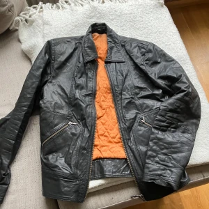 Svart skinnjacka med orange quiltat foder från Nudie jeans vi - Säljer en svart skinnjacka med orange quiltat innerfoder och dragkedja framtill. Jackan har två snedställda fickor med dragkedja och klassisk krage. Perfekt för dig som gillar en stilren och tidlös look.
