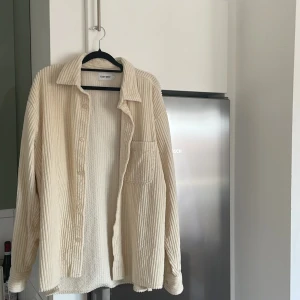 Beige manchester overshirt från East West - Säljer en beige overshirt i manchester från East West. Skjortan har klassisk krage, bröstficka och knäppning framtill. Perfekt att slänga över en t-shirt för en avslappnad look.
