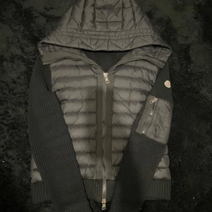 Svart dunjacka med stickade ärmar från Moncler - Säljer en svart dunjacka från Moncler med stickade ärmar och huva. Jackan har dragkedja framtill och på ärmen samt en klassisk Moncler-logga på vänster ärm. Perfekt för kyliga dagar.
