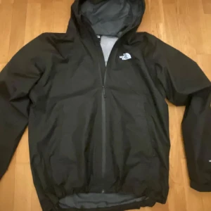 Svart vindjacka från The North Face DryVent - Svart vindjacka från The North Face med DryVent-teknologi. Jackan har huva, dragkedja framtill och meshfoder på insidan. Perfekt för blåsiga dagar och enkel att matcha med allt. Klassisk logga på bröstet.